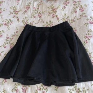 Svart kortkjol från SHEIN - Enkel och snygg svart kortkjol från SHEIN med utsvängd passform. Kjolen är tillverkad i en mjuk blandning av rayon, polyester och spandex som ger en skön stretch. Perfekt att matcha med en crop top eller hoodie för en chill look.Dragkedja i sidan.