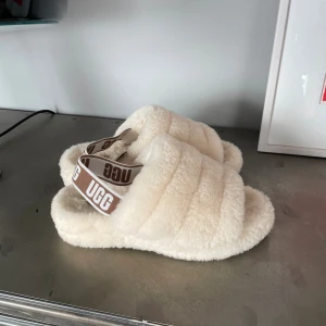 Beige fluffiga sandaler från UGG - Supermysiga sandaler från UGG i beige fluffigt material med breda band och öppen tå. De har en elastisk rem med UGG-logga i brunt och vitt, samt en platt sula i gummi med mönster. Perfekta för chill dagar hemma eller när du vill ha extra komfort.