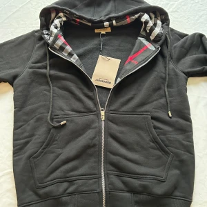 Svart Burberry hoodie med dragkedja - Svart hoodie från Burberry med klassiskt rutigt mönster i huvan och snygga detaljer i rött och grått. Tröjan har huva med snörning, två stora fickor fram och hel dragkedja. Tillverkad i mjuk bomull och polyester för skön komfort.