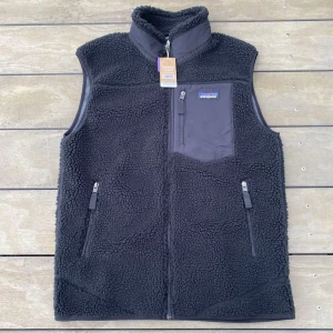 Svart fleeceväst från Patagonia - Mysig svart fleeceväst från Patagonia med hög krage och hel dragkedja. Västen har tre fickor med dragkedja, varav en bröstficka med logga. Perfekt för lager-på-lager och outdoor. Snygg, enkel design med mjukt teddyfoder.