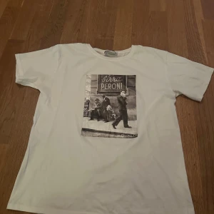 Vit t-shirt med print från Bvaldi - Snygg vit t-shirt från Bvaldi i storlek XS med ett coolt svartvitt print på framsidan som visar en retro stadsscen med texten 'Birra Peroni'. Klassisk rund hals och korta ärmar. Perfekt för dig som gillar streetstyle och grafiska motiv.