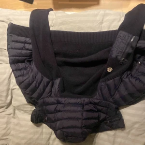 Svart Moncler dunjacka med huva - Säljer en svart dunjacka från Moncler med huva och ribbstickade detaljer. Jackan har dragkedja framtill, quiltad design och en ficka med dragkedja på ärmen. Märket syns tydligt på ärmen och insidan. Perfekt för dig som vill ha en snygg och varm jacka med exklusiv känsla.