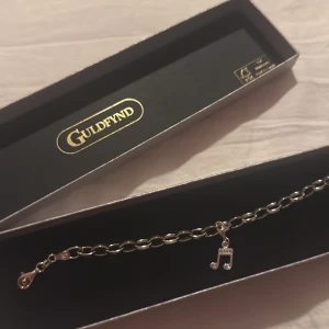 Silverarmband med notberlock från Guldfynd - Stilrent silverarmband från Guldfynd med en kedja i ovala länkar och en hängande berlock formad som en musiknot, dekorerad med små glittriga stenar. Perfekt för dig som älskar musik och vill ha något unikt på handleden.