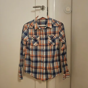 Rutig skjorta från Lee i blå och orange - Säljer en snygg rutig skjorta från Lee i blått, orange och vitt. Skjortan har två bröstfickor med knappar och klassisk krage. Perfekt för dig som gillar avslappnad stil med lite edge.