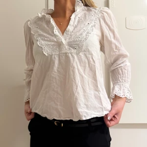 Vit blus med spetsdetaljer - Superfin vit blus i bomull med broderade spetsdetaljer vid krage, bröst och ärmslut. Långärmad modell med lätt v-ringning och volangkant. Perfekt för dig som gillar romantisk och trendig stil.