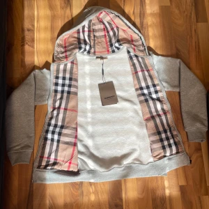 Grå huvtröja från Burberry - OBS: helt oanvänd Säljer en stilren grå huvtröja från Burberry med klassiskt rutigt mönster på insidan av huvan. Tröjan har dragkedja framtill och praktiska fickor. Perfekt för en avslappnad look med en touch av lyx.