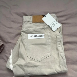 Beige straight jeans från Zara - Beiga raka jeans från Zara, modell 1995 Straight. Klassisk femficksdesign, hög midja och knäppning med flera silverfärgade knappar. Tillverkade i bomull med en mjuk känsla och snyggt läderpatch bak i midjan. Perfekta för en clean och stilren look. Aldrig använda!