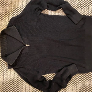 Svart ribbad polotröja med half zip - Svart polotröja i ribbad bomull med half zip-dragkedja vid halsen. Tröjan har lång ärm och en stilren, enkel design som funkar till många olika looks. Materialet är 100% bomull och känns mjukt mot huden. Perfekt för dig som gillar minimalistisk stil.          Jag har tyvärr tagit bort lappen så jag vet inte vilket märke eller storlek de är. Men jag tror jag köpte den på H&M. Den passar folk som är runt 155-175cm långa.