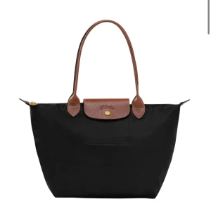 Svart Longchamp Le Pliage handväska - Klassisk svart Le Pliage handväska från Longchamp med bruna skinnhandtag och lock. Väskan har guldfärgad knapp med logga och dragkedja upptill. Tillverkad i slitstark syntet med detaljer i präglat skinn. Perfekt storlek för allt du behöver ha med dig.