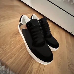 Burberry svarta sneakers med rutig detalj - Snygga svarta sneakers från Burberry med klassisk rutig panel i beige, svart och vit på sidan. Skorna har rund tå, platt vit sula och svarta snören. Insidan är vit och materialet är en mix av skinn och textil. Perfekt för dig som gillar stilrena och lyxiga sneakers.