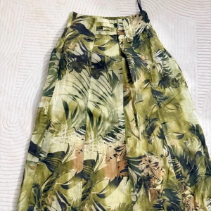 Grön långkjol med tropiskt mönster - Snygg långkjol från atelier GARDEUR i storlek 38. Kjolen har ett tropiskt mönster i gröna, beige och bruna toner med plisserade detaljer. Tillverkad i ett lätt och luftigt material, perfekt för varma dagar.