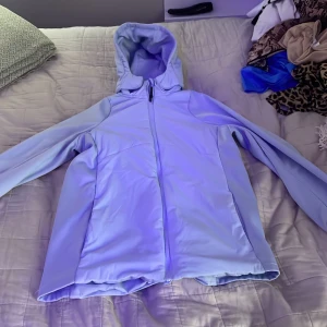 Ljusblå vindjacka med huva - Snygg ljusblå vindjacka med hel dragkedja framtill och praktisk huva. Jackan har långa ärmar med tumhål och släta fickor på sidorna. Materialet är mjukt och känns som syntet, perfekt för blåsiga dagar. Enkel och stilren design som passar till många outfits.