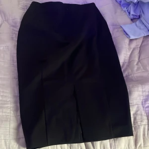 Svart kostymtjol från H&M, storlek 36 - Klassisk svart kostymtjol från H&M i storlek 36. Kjolen har en stilren, figurnära passform med slits framtill och dragkedja i sidan. Perfekt till en snygg topp eller skjorta. Enkel och tidlös design som passar till många olika looks.