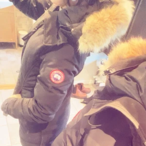 Svart Canada Goose dunjacka med päls - Kan diskutera priserSäljer en svart lång dunjacka från Canada Goose med ikonisk röd logga på ärmen och fluffig beige päls på huvan. Jackan har dragkedja, knappar och flera fickor. Perfekt för kalla dagar och har en snygg, normal passform.