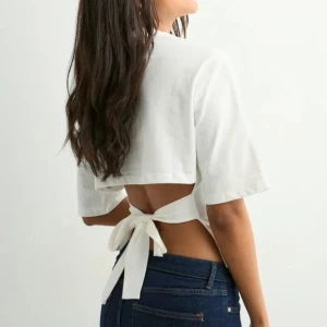 Vit topp med knyt i ryggen - Säljer en vit croppad topp ifrån Nelly med korta ärmar och snygg öppen rygg som knyts med ett brett band. Perfekt för dig som gillar trendiga och unika detaljer. Toppens stilrena look gör den lätt att matcha med dina favoritjeans.
