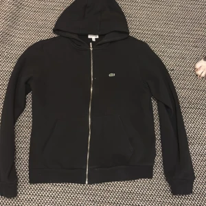 Svart hoodie från Lacoste med dragkedja - Svart äkta  hoodie från Lacoste med klassisk krokodillogga på bröstet. Tröjan har huva, hel dragkedja framtill och två fickor. Materialet är mjuk bomull och passformen är normal. Perfekt för chill dagar och streetwear-stil.