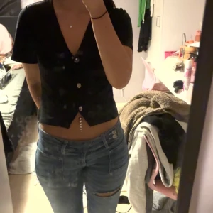 Svart croppad v-ringad topp - Säljer en svart croppad topp med v-ringning och korta ärmar. Toppen har tre dekorativa knappar framtill och sitter snyggt med en slim passform. Perfekt för dig som gillar en enkel men stilren look.