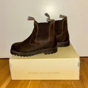 Bruna CRW chelsea boots i läder - Säljer ett par bruna chelsea boots från CRW med grov platt sula och elastiska paneler på sidorna. Skorna har snygga kontrastsömmar och praktiska dragflikar med logga upptill. Tillverkade i slitstarkt läder och har rund tå för en klassisk look.