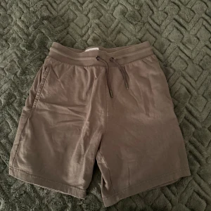 Bruna/ gröna  shorts med snörning från DNMD - Säljer ett par bruna shorts från DNMD i mjuk bomull med elastisk midja och snörning. Shortsen har två sidofickor och en bakficka, perfekt för chill dagar. Klassisk loose fit och enkel design som funkar till det mesta.