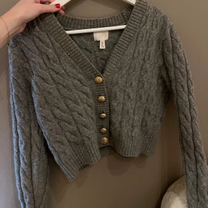 Grå stickad kofta med guldknappar - Superfin grå stickad kofta från H&M med snyggt flätmönster och guldiga knappar framtill. Koftan har v-ringning, ribbade muddar och lång ärm. Perfekt att slänga över en topp för en chill look.