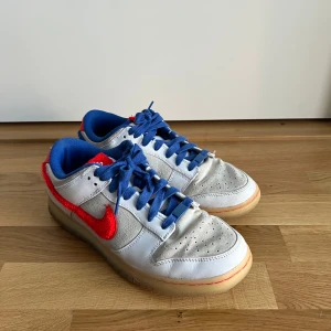 Nike Dunk Low vit/blå/röd - Nike Dunk Low year of the rabbit 2023 limited edition sneakers med vit och grå ovandel i skinn och mocka, blå meshfoder och snörning samt röd, fluffig Swoosh och häl. Yttersulan är gummifärgad och innersulan har ett rött grafiskt tryck. Snygg kontrast mellan färgerna och coola detaljer.