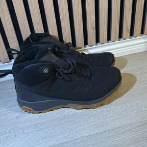 Svarta vattentäta kängor från Salomon - Säljer ett par svarta kängor från Salomon med grov brun sula och snörning. Perfekta för kommande vintern. Skorna har en robust design med högt skaft och förstärkta partier i syntetmaterial, perfekta för utomhusaktiviteter. 
