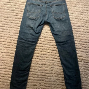 Blå tapered jeans från Jack and jones - Snygga blå jeans från Jack and jones i modellen slim glenn, Jeansen har en klassisk femficksdesign och smal passform. Jeansen har en mörk tvätt och raka ben med lätt slitning framtill. Perfekta för dig som gillar en modern och stilren look.