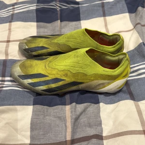 Gula fotbollsskor från Adidas - Säljer ett par gula fotbollsskor från Adidas med svarta ränder på sidan. Skorna har en strumpliknande ovandel i syntetmaterial och en platt sula med dobbar för bra grepp på planen. De har en modern, strömlinjeformad design utan snörning och är perfekta för dig som vill sticka ut.