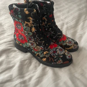 Blommiga svarta kängor med snörning - Unika svarta kängor med färgglatt blommigt mönster i rött, vitt, grönt och gult. Skorna har rund tå, platt sula och snörning framtill. Materialet är troligtvis syntet med textilkänsla. Perfekt för dig som vill sticka ut med din stil. Aldrig använda endast testade! 