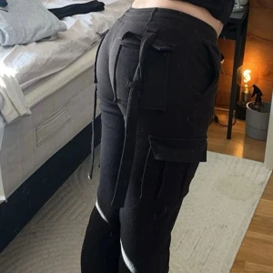 Svarta cargopants - Svarta cargopants med coola fickor på sidorna och snörningsdetaljer både fram och bak. Byxorna har en avslappnad passform och är tillverkade i mjukt bomullsmaterial. Midjan är låg, och har sytt om dem för att få den klassiskt lägmidjade passformen. 