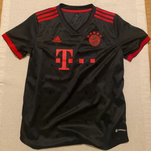 FC Bayern München t-shirt - Snygg svart FC Bayern München fotbollströja med röda detaljer och tryck, från deras 2022/2023 säsong. Inte använd mer än att jag testat den annars har den bara legat i garderoben så den är som ny