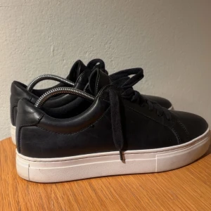 Svarta sneakers från Vagabond - Svarta sneakers från Vagabond i skinn med klassisk design och vita platta sulor. Skorna har rund tå, svarta snören och stilren look som passar till det mesta. Perfekta för dig som gillar enkel och clean stil Dem är i storlek 39! inga defekter och jättebra skötsamma, nästan i nyttskick. Pris inte fast och kan diskuteras!