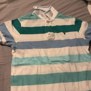 Randig pikétröja från Ralph Lauren - Snygg pikétröja från Ralph Lauren med breda ränder i vitt, blått och grönt. Klassisk krage med knappar och den ikoniska broderade polospelaren på bröstet. Perfekt för dig som gillar sportig och avslappnad stil.