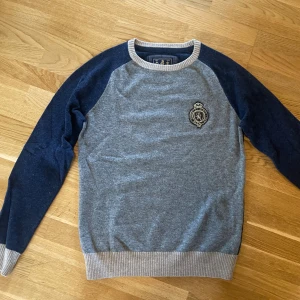 Grå och marinblå merinoullströja Morris - Snygg tröja från Morris i 100% merinoull, grå framtill och marinblå ärmar med kontrasterande beige muddar. Broderad logga på bröstet och coola mockadetaljer på armbågarna. Perfekt för dig som gillar klassisk men trendig stil.