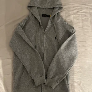 Grå Ralph Lauren zip - Snygg grå Ralph Lauren med dragkedja och huva. Klassisk broderad logga på bröstet och ribbade muddar. Väldigt lite använd, inga skador eller fläckar. Säljer den på grund av att den är lite för kort i armarna på mig. Jag är 178cm, perfekt i längd med som sagt kort i armar, skulle därför säga att den sitter som M/S.