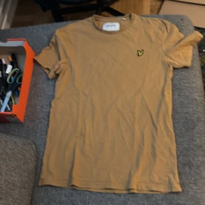 Beige t-shirt från Lyle & Scott - Snygg beige t-shirt från Lyle & Scott med klassisk rund hals och korta ärmar. Gjord i mjuk ekologisk bomull och har den ikoniska broderade gula örnen på bröstet. Perfekt för en chill och stilren look.