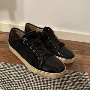 svarta lanvins  - Snygga svarta sneakers från Lanvin med klassisk snörning och rund tå. Ovandelen är i mocka och tån är i blankt lackskinn, vilket ger en cool kontrast. Sulan är beige och platt, och insidan är fodrad i skinn. Perfekta för dig som gillar stilrena detaljer.