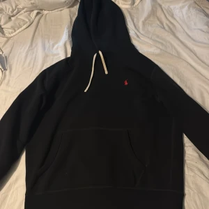 Svart hoodie från Ralph Lauren - Svart hoodie från Ralph Lauren med klassisk röd logga broderad på bröstet. Tröjan har vit snörning i huvan och en stor magficka framtill. Perfekt för en chill och stilren look.