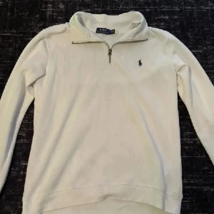 Vit half zip tröja från Polo Ralph Lauren - Vit långärmad tröja från Polo Ralph Lauren med klassisk half zip-dragkedja och broderad logga på bröstet. Tröjan har en stilren design och är tillverkad i mjuk bomull, perfekt för lager-på-lager under kyliga dagar.