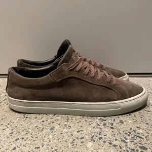 Bruna mockasneakers med vit sula - Snygga bruna sneakers i mocka med rund tå och platt vit sula. Skorna har matchande bruna snören och en stilren, minimalistisk design. Insidan är svart och de passar perfekt för dig som gillar en clean och klassisk look.de 