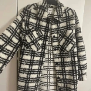 Rutig teddyjacka från Kappahl - Säljer en oversized teddyjacka från Kappahl i svartvitt rutigt mönster. Jackan har stora knappar framtill, klassisk krage och två bröstfickor. Materialet är fluffigt och mjukt, perfekt för kyliga dagar. Jackan har lång ärm och en riktigt trendig vibe.