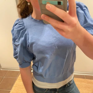 Blå topp med puffärm från Lindex - Säljer en ljusblå topp från Lindex med korta puffärmar och rund halsringning. Toppen har ribbade muddar vid ärmslut och midja, vilket ger en snygg passform. Perfekt för dig som gillar en trendig och söt stil.