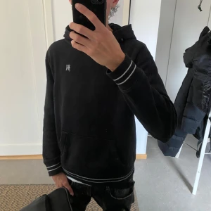 Svart hoddie från hollister - Svart hoodie från Hollister med vit broderad logga på bröstet och tryck på huvan. Tröjan har vita ränder på muddar och nederkant, känguruficka och huva. Perfekt till vintern. Han på bilden är 187.