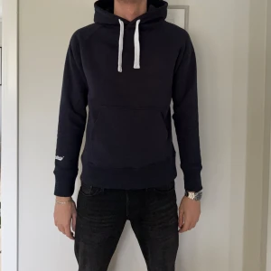 Blå hoodie från Russemerch med huva - Nypris 499kr, denna är helt oanvänd! Blå hoodie från Russemerch med vit snörning i huvan och klassisk känguruficka framtill. Tröjan har lång ärm och en stilren look, perfekt för dig som gillar enkel och clean streetstyle. Mjuk bomullskänsla och relaxed passform.