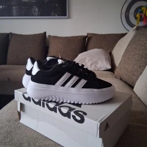 Svarta och vita sneakers från Adidas - Helt nya Snygga Adidas sneakers inte använda en enda gång. i svart och vitt med klassiska tre ränder på sidan. Skorna har en tjock, vit plattformssula och snörning framtill. Ovandelen är i syntet och mocka, med rund tå och Adidas-logga på plösen och hälen. Perfekt för dig som gillar sportig stil. Helt nya a