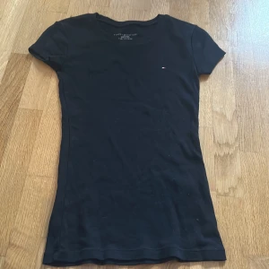 Svart t-shirt från Tommy Hilfiger - Säljer en svart t-shirt från Tommy Hilfiger i slim fit med korta ärmar och rund hals. Diskret Hilfiger-logga broderad på bröstet. Mjuk och skön bomull som sitter snyggt och passar perfekt till jeans eller kjol. Enkelt och stilrent plagg för dig som gillar klassisk design.