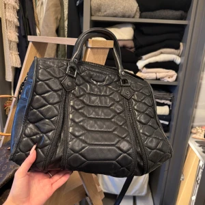 Svart quiltad handväska Zadig & Voltaire - Cool svart quiltad handväska från Zadig & Voltaire i mjukt skinn. Väskan har dubbla handtag, avtagbar axelrem och snygga dragkedjedetaljer framtill. Rymligt innerfack med extra ficka och logga på remmen. Perfekt för dig som gillar edgy och stilren design.