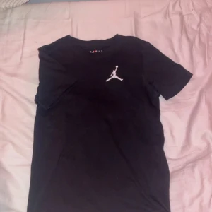 Svart Jordan t-shirt med logga - Svart t-shirt från Jordan med klassisk Jumpman-logga broderad i vitt på bröstet. T-shirten har rund hals, korta ärmar och är gjord i mjuk bomull. Enkel och stilren design som passar perfekt till streetwear.