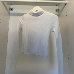Vit ribbad polotröja - Säljer en vit ribbad polotröja med lång ärm och hög krage. Tröjan är figurnära och har en lätt vågig kant nedtill. Perfekt att styla med jeans eller kjol för en clean look.