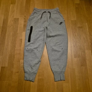 Grå mjukisbyxor från Nike, storlek S - Grå mjukisbyxor från Nike med svarta detaljer och dragsko i midjan. Byxorna har ribbade muddar vid bensluten, en svart dragkedjeficka på ena benet och Nike-logga på höften. Perfekta för chill eller träning.